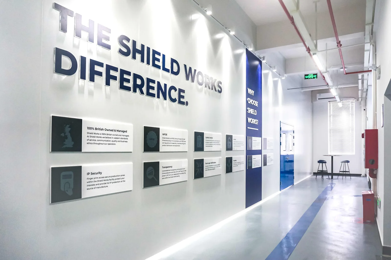 The Shield Works Difference wall running down a corridor inside the Zhuhai facility, with plaques for British ownership, WFOE status, quality control, access, IP security, transparency, dual-layer verification and flexibility
